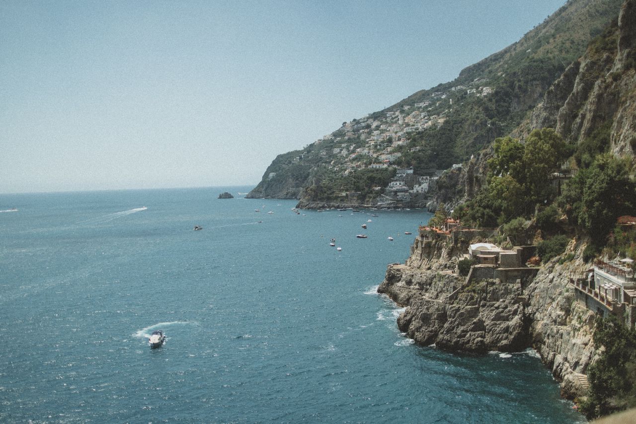 our-story Stunning aerial view of Furore, Italy, showcasing the coastline and Tyrrhenian Sea under a clear blue sky.